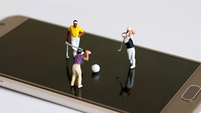 Three miniature figures of golfers posed on the screen of a smartphone. Two of the golfers are poised to tee off. Social media concept, smart technology, club marketing.
